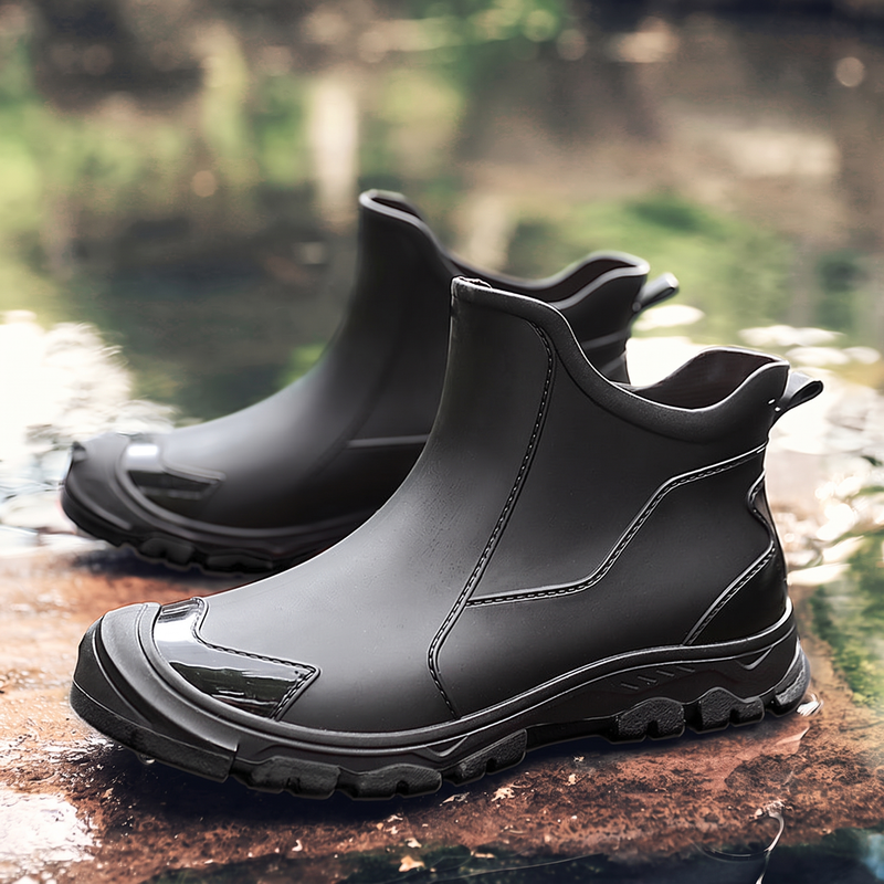 A pair of black rubber ankle boots with a side logo patch, shown on a rock near water.