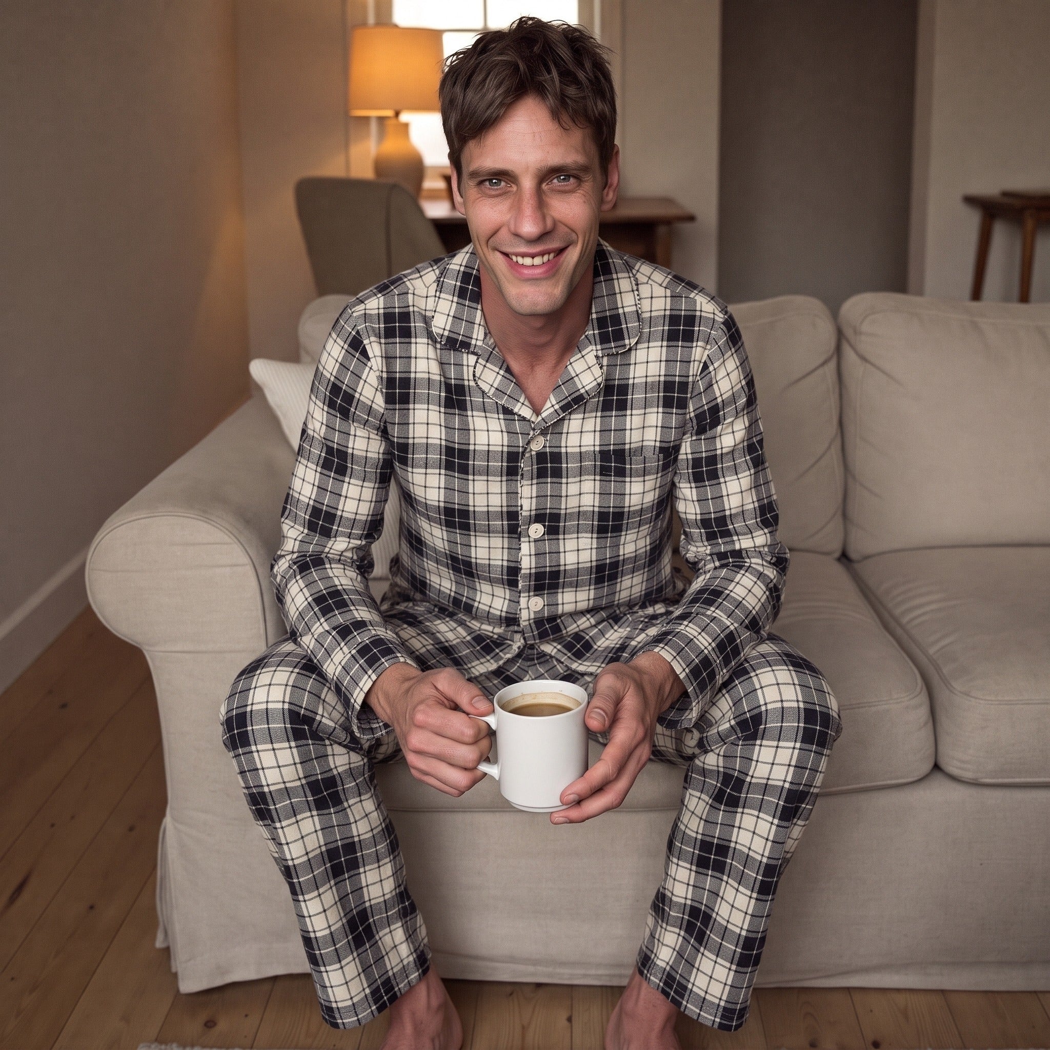 Man wearing navy and cream check flannel pyjamas, sitting on sofa holding mug | Chaps London