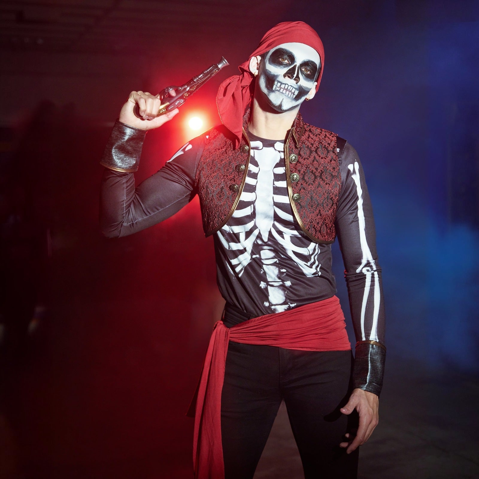 Man wearing skeleton shirt, red waistcoat, headscarf and sash, holding prop gun | Chaps London