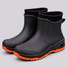 Black ankle rain boots with orange lugged soles and black ribbed knit cuffs, three quarter view on a grey studio background.