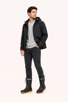 Man in a black hooded jacket, grey t-shirt and dark jeans wearing black mid calf rain boots with orange soles, a reflective band and drawstring tops.